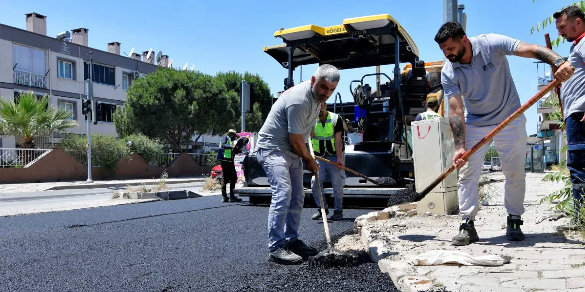 Bornova Belediyesi ekipleri, bir sokakta asfalt serim çalışması yapıyor. İşçiler, ellerindeki tırmık ve küreklerle yeni dökülen sıcak asfaltı düzeltirken, arkalarında sarı renkli bir asfalt serme makinesi bulunuyor. Arka planda ise çok katlı konutlar ve ağaçlar yer alıyor.