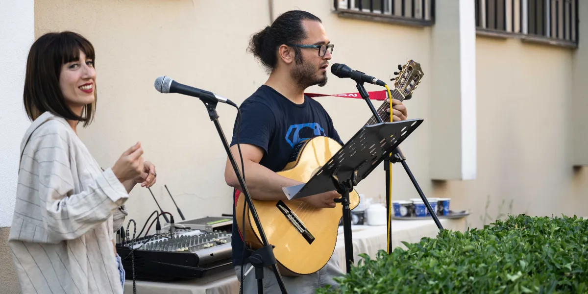 Açık havada, bir binanın önünde canlı müzik performansı sergileyen iki kişi. Gözlüklü, saçları arkadan bağlı bir erkek, üzerinde Superman logolu tişörtüyle akustik gitar çalıp mikrofon başında şarkı söylüyor. Yanında ise açık renkli, çizgili bir ceket giyen kadın, ritim tutarak eşlik ediyor. Önlerinde bir ses mikseri ve nota sehpası bulunuyor.