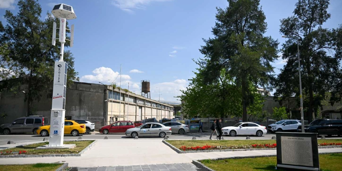 Bornova'da bir cadde kenarında, üzerinde güneş panelleri ve dikey rüzgar türbini bulunan, Rumitech logolu modern bir aydınlatma direği yer alıyor. Direğin hemen yanında, üzerinde 