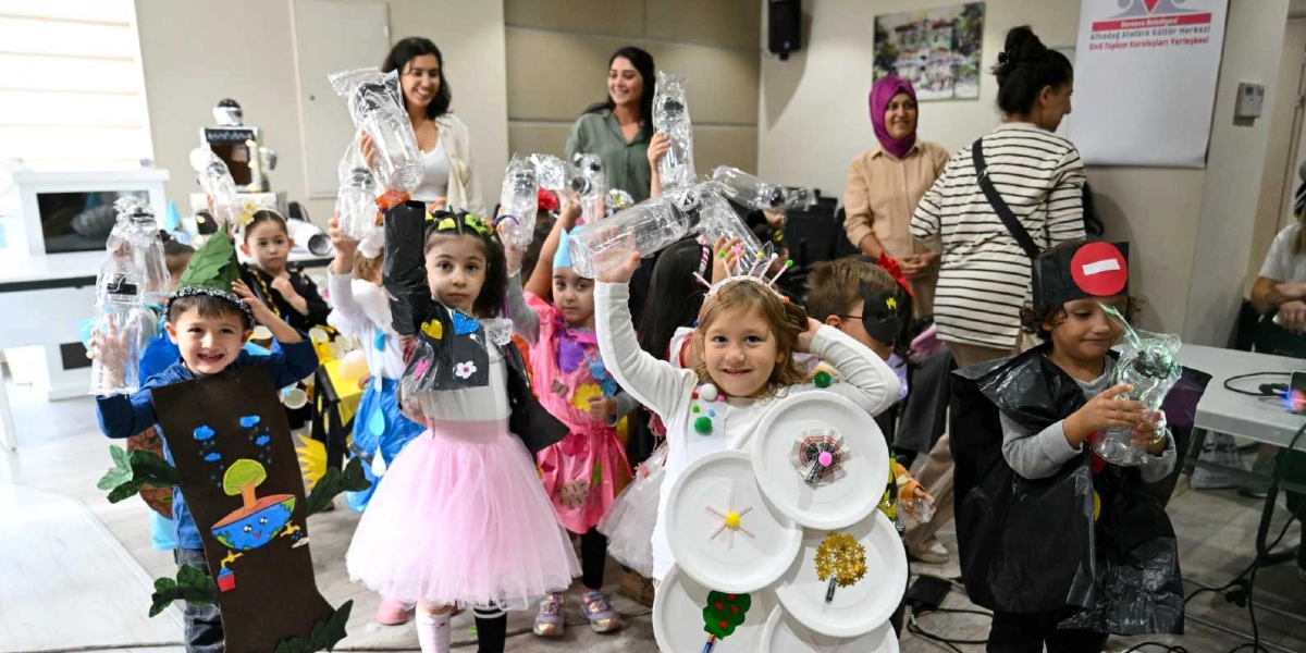 Geri dönüştürülebilir atıklardan yapılmış kostümler giyen bir grup çocuk, ellerinde plastik şişelerle poz veriyor. Çocuklardan biri ağaç figürlü bir kıyafet, diğeri beyaz tabaklardan yapılmış bir kostüm, bir diğeri ise siyah poşetten yapılmış bir pelerin giyiyor. Arka planda çocuklara eşlik eden yetişkinler ve duvarda Bornova Belediyesi Altındağ Atatürk Kültür Merkezi'ne ait bir tabela bulunuyor.