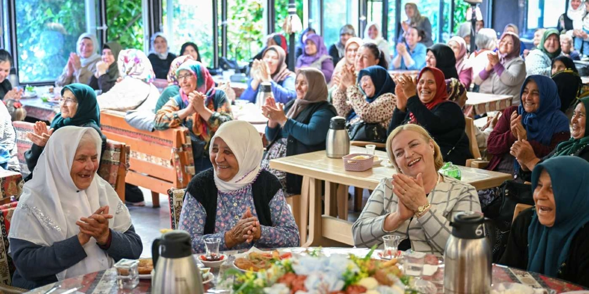 Kapalı bir mekanda, Anneler Günü kutlaması için bir araya gelmiş, çoğu başörtülü olan neşeli bir kadın grubu. Masalarda oturan kadınlar gülümseyerek alkış tutuyor, önlerindeki masalarda çay bardakları ve yiyecekler bulunuyor. Ortamda samimi ve dayanışma dolu bir atmosfer hakim.
