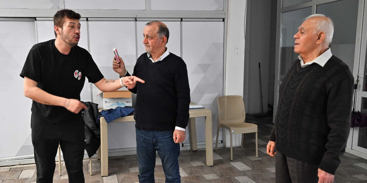 İç mekan bir alanda, siyah tişörtlü genç bir eğitmen, elinde küçük bir nesne tutan orta yaşlı bir adam ve onu dinleyen yaşlı bir adam yaratıcı drama çalışması yapıyor. Eğitmen, elini uzatarak diğer katılımcıya bir şeyler anlatırken, katılımcılar dikkatle onu takip ediyor. Arka planda beyaz bir masa, sandalyeler ve cam bölmeli bir duvar bulunuyor.