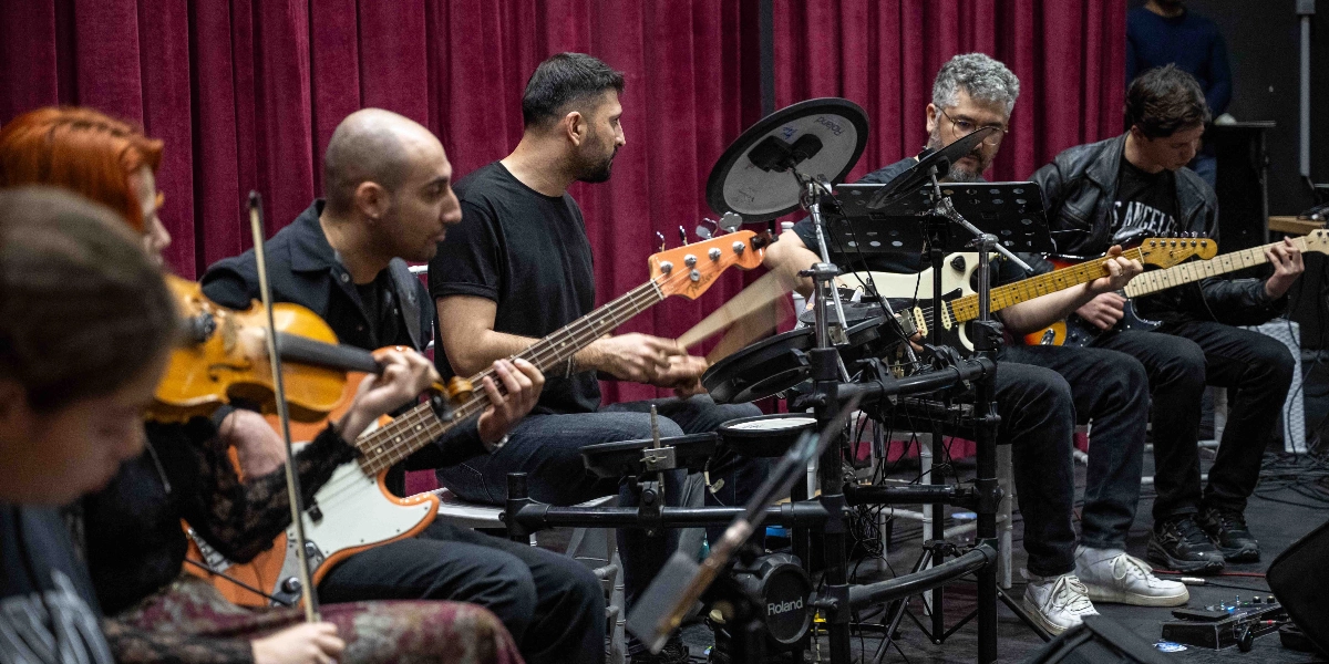 Kırmızı perdeli bir sahnede prova yapan müzik grubu; ön planda keman çalan bir kadın, yanında bas gitar çalan bir erkek, ortada elektronik bateri çalan bir müzisyen, sağ tarafta ise elektro gitar çalan iki erkek müzisyen yer alıyor.