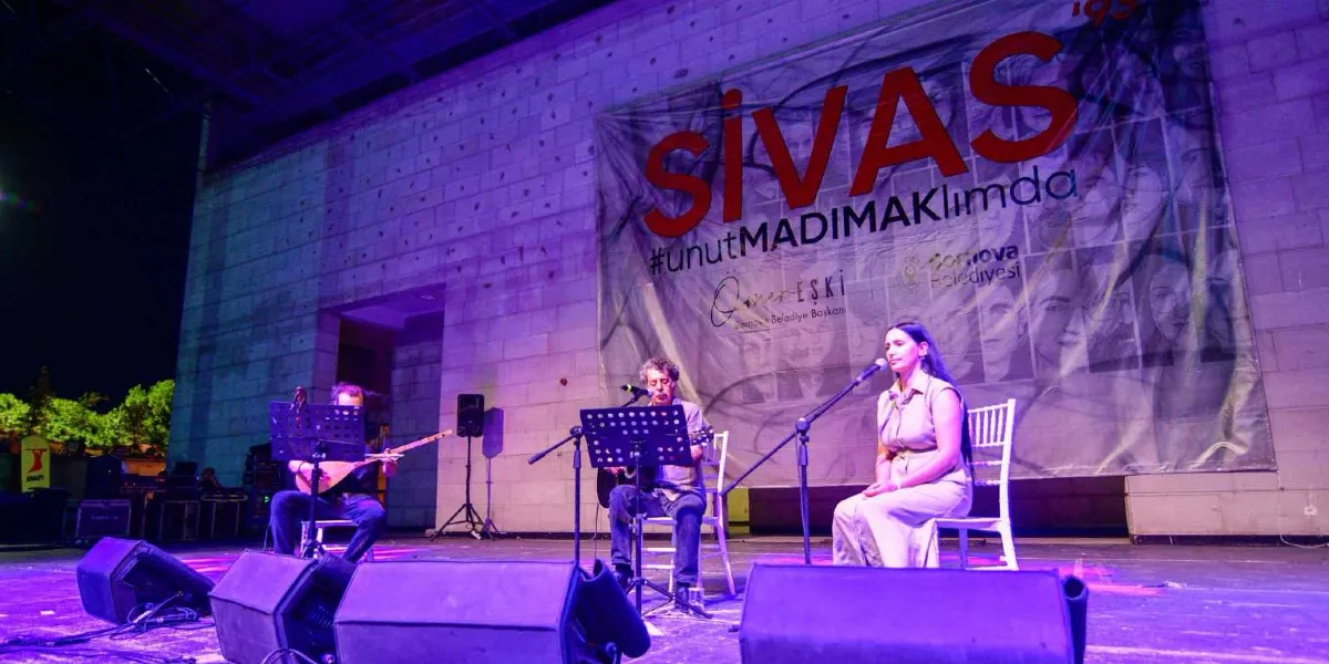 Sivas Madımak anma etkinliğinde, sahnede oturan üç müzisyen performans sergiliyor. Solda bağlama çalan bir erkek, ortada gitar çalan bir erkek ve sağda şarkı söyleyen bir kadın bulunuyor. Arka planda, üzerinde 