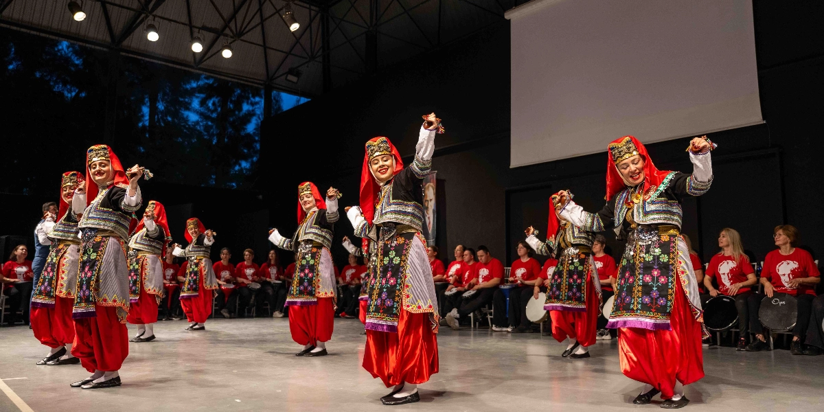 Geleneksel Türk halk dansları kıyafetleri giymiş bir grup kadın, kapalı bir sahnede dans ediyor. Dansçılar, kırmızı şalvarları, işlemeli yelekleri ve başlarında altın süslemeli kırmızı başlıklarıyla kollarını havaya kaldırmış, ellerinde zillerle figür sergiliyorlar. Sahnenin arka planında oturan ve üzerinde Atatürk baskılı kırmızı tişörtler giyen bir müzik grubu, ellerindeki davullarla ritim tutuyor. Sahne ışıklarıyla aydınlatılan ortamda, arkada büyük bir projeksiyon perdesi ve ağaçların göründüğü karanlık bir dış mekan bulunuyor.