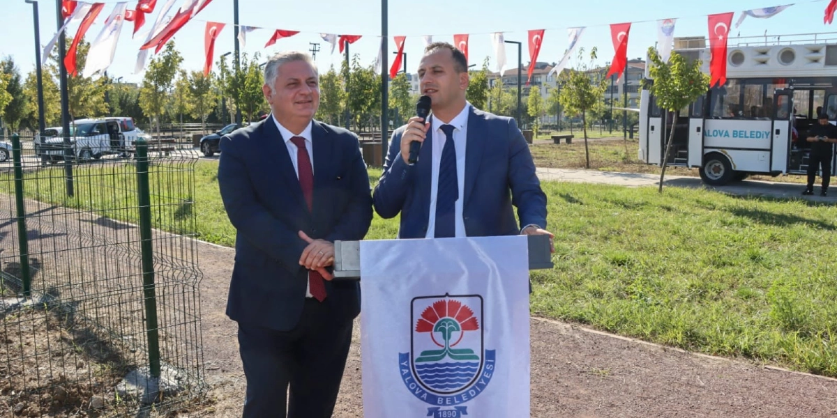 Açık havada, üzerinde Yalova Belediyesi logosu bulunan bir kürsüde konuşma yapan takım elbiseli bir erkek ve yanında duran diğer takım elbiseli bir erkek. Arka planda Türk bayraklarıyla süslenmiş bir alan ve üzerinde Yalova Belediyesi yazan bir otobüs bulunuyor.