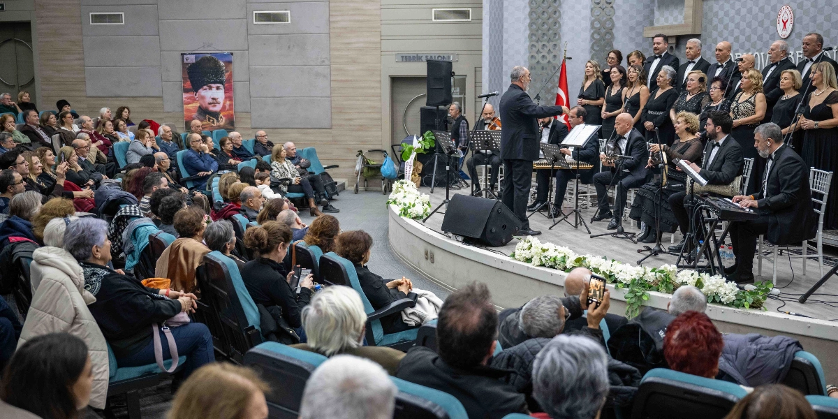 Modern bir konferans salonunda, sahnede siyah kıyafetler giymiş bir koro ve orkestra müzik icra ediyor. Şef, orkestrayı yönetirken, izleyiciler koltuklarda oturarak konseri takip ediyor. Salonun duvarında Mustafa Kemal Atatürk'ün büyük bir portresi asılı duruyor ve sahne önü beyaz çiçeklerle süslenmiş durumda. İzleyicilerin bir kısmı telefonlarıyla çekim yapıyor.