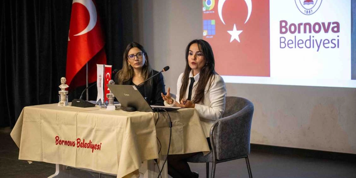Bir sunum sırasında masada yan yana oturan iki kadın konuşmacı. Sağ taraftaki kadın beyaz ceketli, el hareketleriyle bir şeyler anlatıyor; sol taraftaki kadın ise siyah ceketli ve gözlüklü, onu dinliyor. Masanın üzerinde dizüstü bilgisayar, mikrofonlar ve küçük bir Atatürk büstü bulunuyor. Masanın ön yüzünde kırmızı renkle 