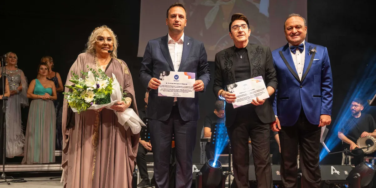 Sahne üzerinde dört kişi yan yana durarak ellerinde plaket veya teşekkür belgesi tutuyor. En solda, elinde büyük bir çiçek buketi tutan sarı saçlı bir kadın, yanında koyu renk takım elbiseli bir erkek, onun yanında siyah işlemeli ceket giyen bir erkek ve en sağda mavi ceketli, papyonlu bir erkek yer alıyor. Arka planda bir müzik grubu ve sahne ışıkları görülüyor.