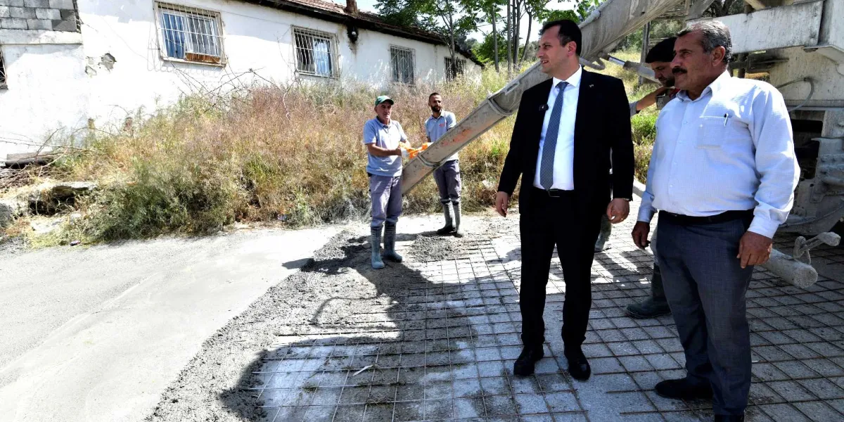 Bornova Belediye Başkanı, takım elbiseli ve kravatlı bir şekilde, yol yapım çalışmalarının devam ettiği bir alanda incelemelerde bulunuyor. Arka planda beton mikserinden dökülen harcı yönlendiren işçiler ve yan tarafta duran bir yetkili yer alıyor. Çalışma alanında zemin üzerine yerleştirilmiş çelik hasır demirler ve dökülmeye başlanan beton görülüyor.