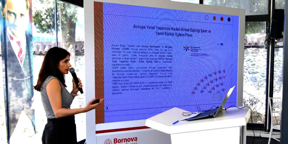 Koyu saçlı bir kadın, elinde mikrofonla Bornova Belediyesi logolu bir sunum ekranının önünde konuşma yapıyor. Ekranda 