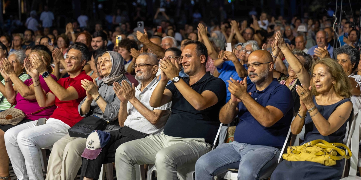 Açık havada düzenlenen bir etkinlikte, sandalyelerde yan yana oturan ve alkışlayarak konseri izleyen kalabalık bir izleyici grubu. Ön planda, farklı yaş gruplarından kadın ve erkeklerin neşeli bir ifadeyle sahneye doğru baktığı, bazılarının ellerinde telefonlarıyla çekim yaptığı görülüyor.