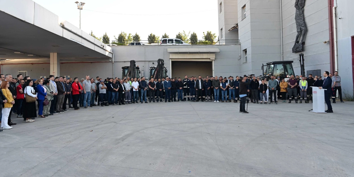 Bornova Belediyesi şantiye alanında, bir kürsüde konuşma yapan yetkiliyi dinleyen kalabalık bir çalışan grubu. Arka planda iş makineleri ve belediye binasının duvarında yer alan bir kabartma heykel bulunuyor.