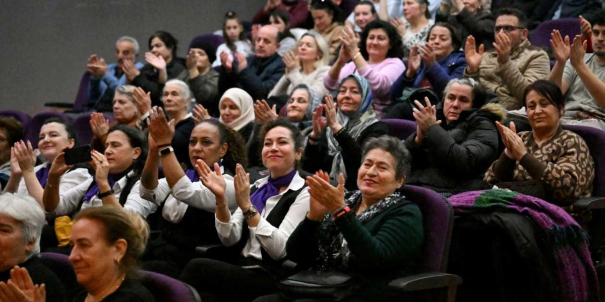 Kapalı bir salonda, mor koltuklarda oturan ve sahnedeki bir etkinliği alkışlayan, ağırlıklı olarak kadınlardan oluşan kalabalık bir izleyici grubu. Ön sıralarda beyaz gömlek üzerine mor fular takmış kadınlar dikkat çekerken, izleyicilerin bir kısmı gülümseyerek sahneye bakıyor, bazıları ise cep telefonlarıyla çekim yapıyor.