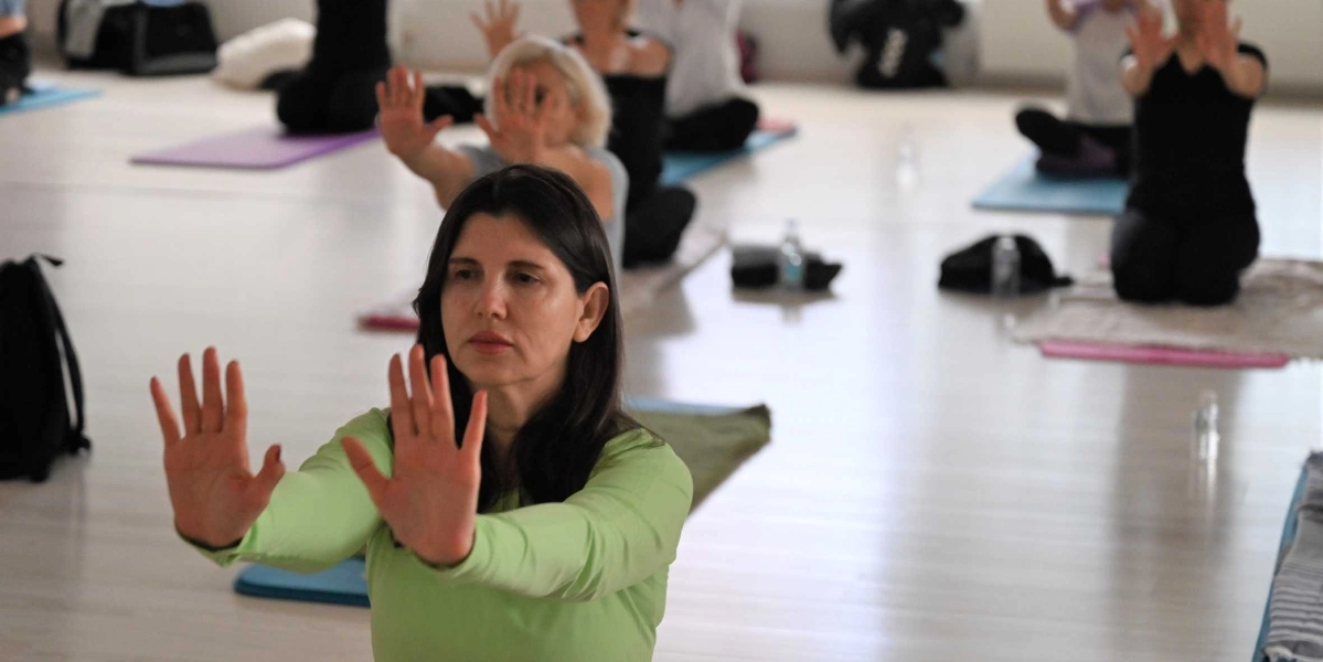 Aydınlık bir stüdyoda yoga matları üzerinde oturan bir grup kadın, ellerini ileriye doğru açarak odaklanmış bir şekilde yoga hareketi yapıyor. Ön planda, açık yeşil uzun kollu bir tişört giyen kadın, elleri karşıya bakacak şekilde kollarını uzatmış, ciddi ve huzurlu bir ifadeyle ileriye doğru bakıyor. Arka planda ise aynı hareketi yapan diğer katılımcılar bulanık bir şekilde görülüyor.