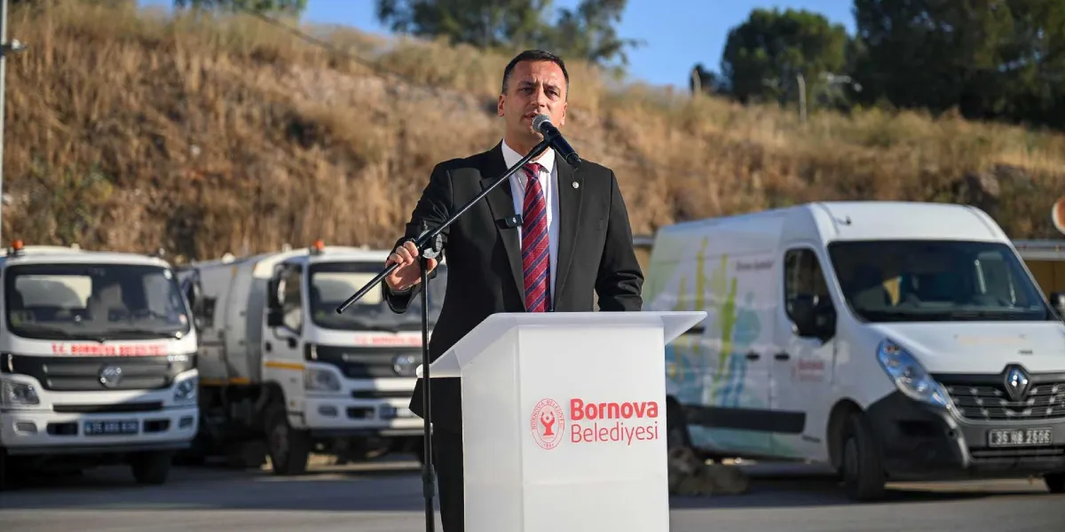 Siyah takım elbiseli ve kırmızı çizgili kravatlı bir erkek, üzerinde Bornova Belediyesi logosu bulunan bir kürsüde mikrofonla konuşma yapıyor. Arka planda, üzerinde Bornova Belediyesi yazılı beyaz hizmet araçları ve toprak bir yamaç bulunuyor.