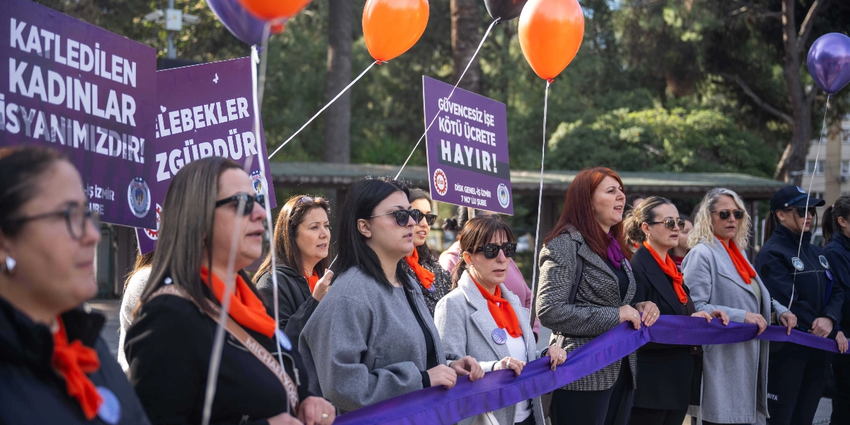 Bornova'da düzenlenen 25 Kasım Kadına Yönelik Şiddetle Mücadele Günü eyleminde, ellerinde mor bir şerit tutan ve boyunlarında turuncu fularlar takan bir grup kadın yan yana duruyor. Kadınların arkasında 