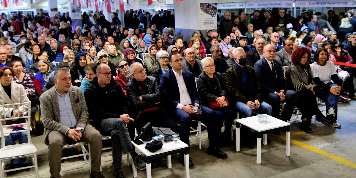 Kapalı bir alanda düzenlenen Tohum Takas Şenliği'nde, sandalyelerde yan yana oturarak etkinliği izleyen kalabalık bir grup. Ön sırada, aralarında Bornova Belediye Başkanı Mustafa İduğ'un da bulunduğu, resmi ve günlük kıyafetli kadın ve erkeklerden oluşan protokol ve katılımcılar yer alıyor. Arka planda ise stantların bulunduğu, farklı yaş gruplarından insanların katıldığı oldukça yoğun bir izleyici kitlesi görülüyor. Salonun tavanında asılı Türk bayrakları ve çeşitli kurumların tabelaları dikkat çekiyor.