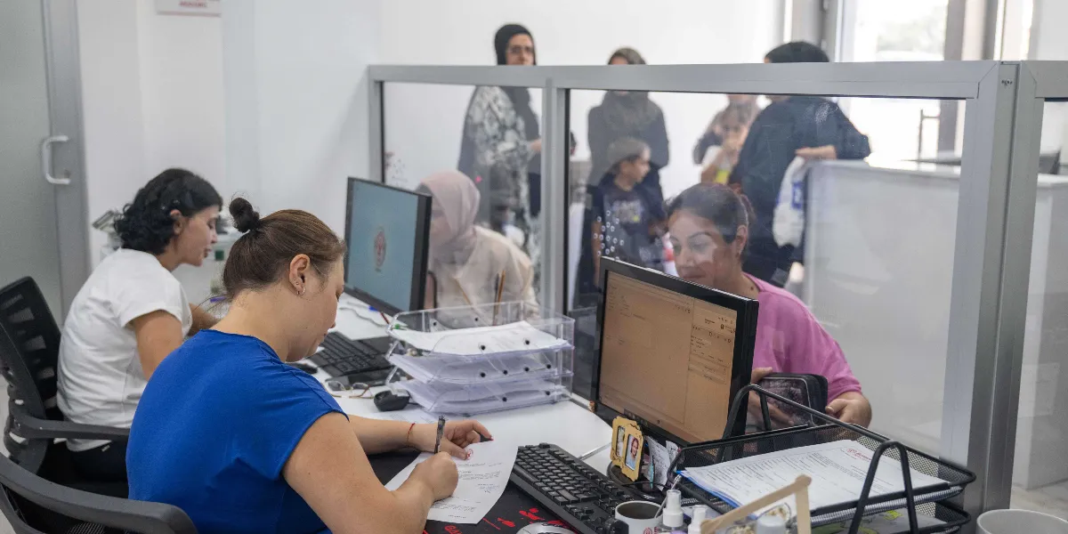 Ofis ortamında, cam bölmelerle ayrılmış masalarda çalışan görevliler ve işlem yapmak için bekleyen vatandaşlar görülüyor. Mavi tişörtlü bir kadın görevli masasında evrak doldururken, yanında beyaz tişörtlü bir çalışma arkadaşı ve karşı tarafta pembe tişörtlü başka bir görevli bilgisayar başında hizmet veriyor. Arka planda ise başvuru yapmak için bekleyen kadınlar ve çocuklar bulunuyor.