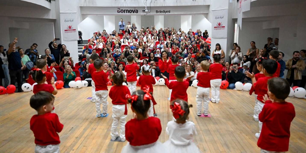 Bornova Belediyesi'ne ait bir salonda, kırmızı üstler ve beyaz altlar giymiş bir grup küçük çocuk, izleyicilere dönük şekilde sahnede gösteri yapıyor. Çocukların saçlarında kırmızı kurdeleler bulunuyor ve yerlerde kırmızı beyaz balonlar yer alıyor. Sahnenin arkasındaki tribünlerde oturan kalabalık bir izleyici grubu, çocukları alkışlayarak izliyor.