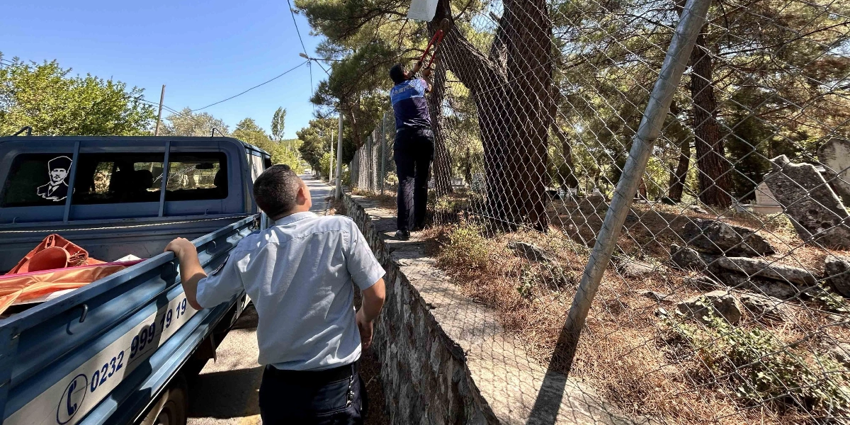 Bornova Belediyesi zabıta ekipleri, yol kenarındaki tel çitlere asılmış olan izinsiz reklam afişlerini büyük bir makas yardımıyla keserek söküyor. Mavi renkli zabıta kamyonunun yanında duran bir görevli çalışmayı izlerken, diğer görevli çitlerin üzerindeki afişi temizliyor. Kamyonun arka camında Atatürk silüeti ve üzerinde telefon numarası bulunan bir çıkartma yer alıyor.
