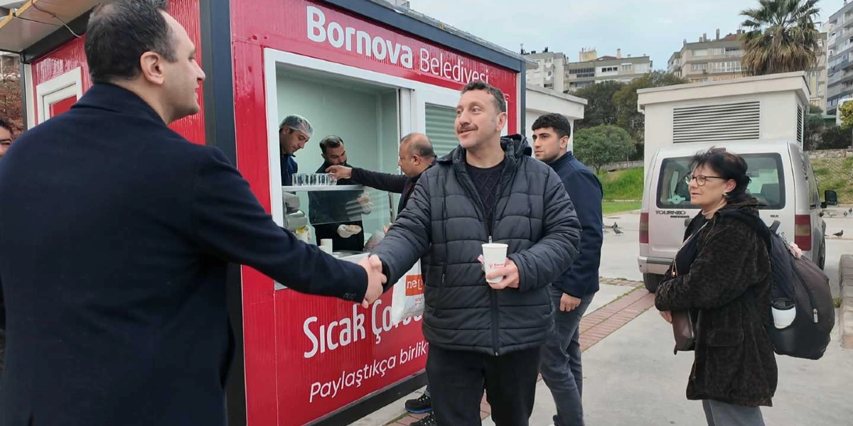 Bornova Belediyesi'ne ait kırmızı renkli bir mobil ikram aracının önünde, lacivert ceketli bir erkek, elinde karton bardak tutan siyah şişme montlu bir erkekle tokalaşıyor. Arka planda belediye personeli ikram hazırlığı yaparken, yan tarafta gözlüklü bir kadın ve genç bir erkek onları izliyor. Aracın üzerinde 