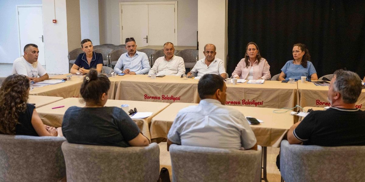 U-şeklinde birleştirilmiş masalar etrafında toplanmış bir grup insan toplantı yapıyor. Masaların üzerinde Bornova Belediyesi yazılı örtüler serili. Arka tarafta yan yana oturan altı kişi ve ön tarafta sırtları dönük oturan üç kişi bulunuyor. Katılımcılar resmi olmayan bir ortamda, masadaki notlar ve belgeler eşliğinde görüşme gerçekleştiriyor.