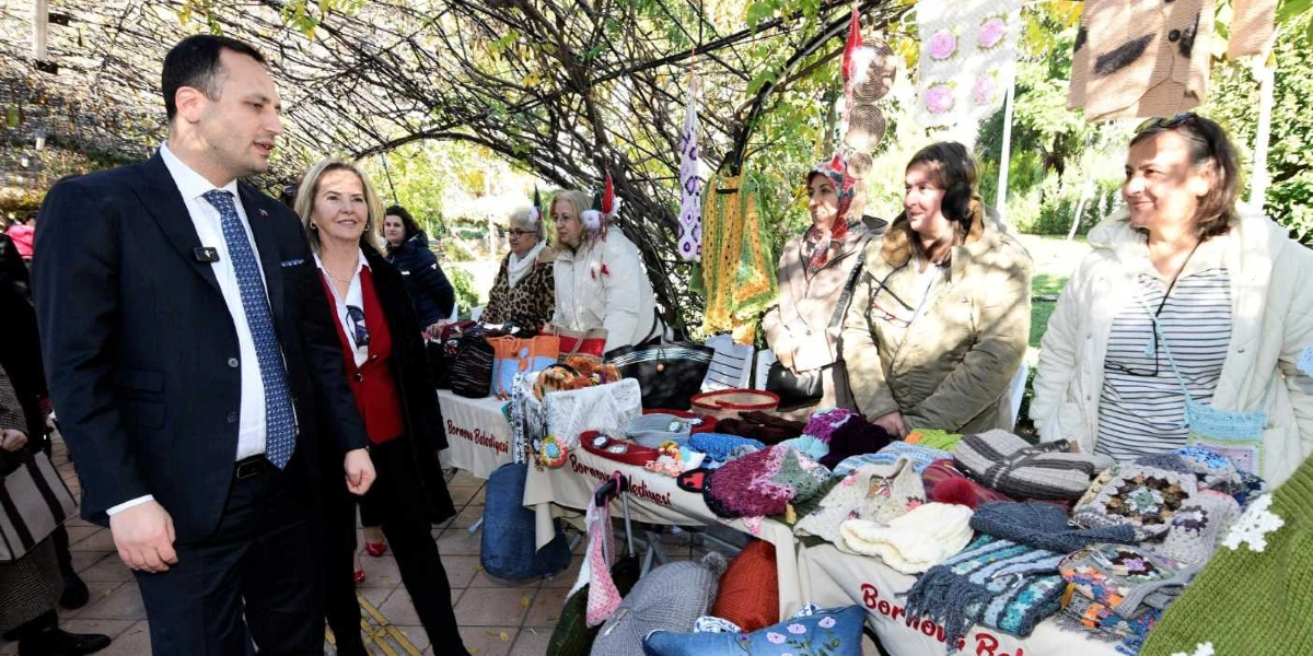 Açık havada kurulan bir stantta, Bornova Belediyesi logolu örtülerin üzerinde sergilenen el yapımı örgü ürünleri, çantalar ve hediyelik eşyaları inceleyen bir grup insan yer alıyor. Lacivert takım elbiseli bir erkek ve yanında kırmızı ceketli bir kadın, stanttaki ürünleri incelemek için durmuş, stant görevlisi kadınlarla sohbet ediyor. Arka planda ağaçların altında, kışlık kıyafetler giymiş diğer katılımcılar ve sergilenen ürünler görülüyor.