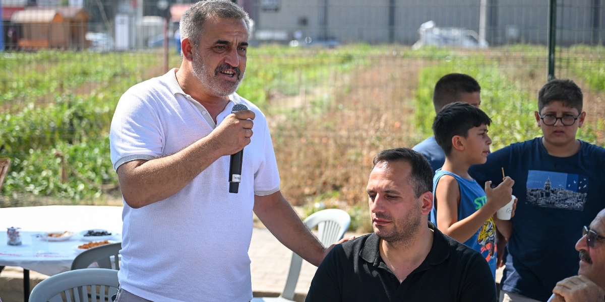 Beyaz polo yaka tişörtlü, orta yaşlı bir adam elinde mikrofonla konuşma yaparken, yanında siyah tişörtlü bir adam oturuyor. Arka planda ise bir grup çocuk ve tarım arazisi bulunuyor.