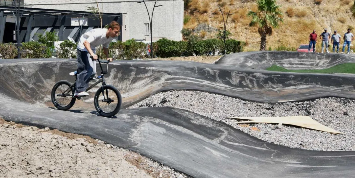Açık havada, engebeli ve kıvrımlı asfalt bir parkurda BMX bisikleti süren genç bir erkek. Arka planda parkuru izleyen dört kişilik bir grup insan, ağaçlar ve modern bir yapı bulunuyor.