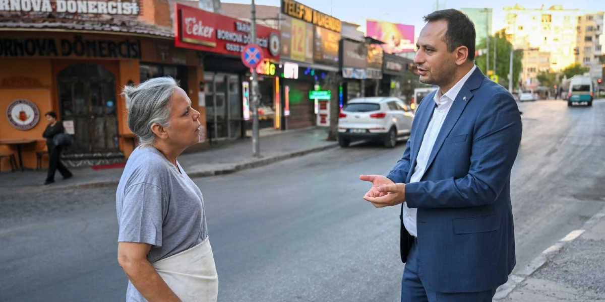 Bornova'da bir cadde üzerinde, gri tişörtlü ve beyaz önlüklü bir kadın ile lacivert takım elbiseli bir erkek ayakta sohbet ediyor. Arka planda dükkanlar, park halindeki araçlar ve sabah saatlerinin sakinliği görülüyor.