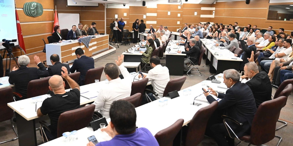 Bornova Belediyesi meclis salonunda düzenlenen toplantıda, katılımcıların büyük çoğunluğu ellerini havaya kaldırarak oylama yapıyor. Salonun ön kısmındaki başkanlık divanında yetkililer otururken, izleyici ve meclis üyeleri sıralarda yer alıyor. Arka duvarda Atatürk rölyefi, Türk bayrağı ve Bornova Belediyesi flaması bulunuyor.