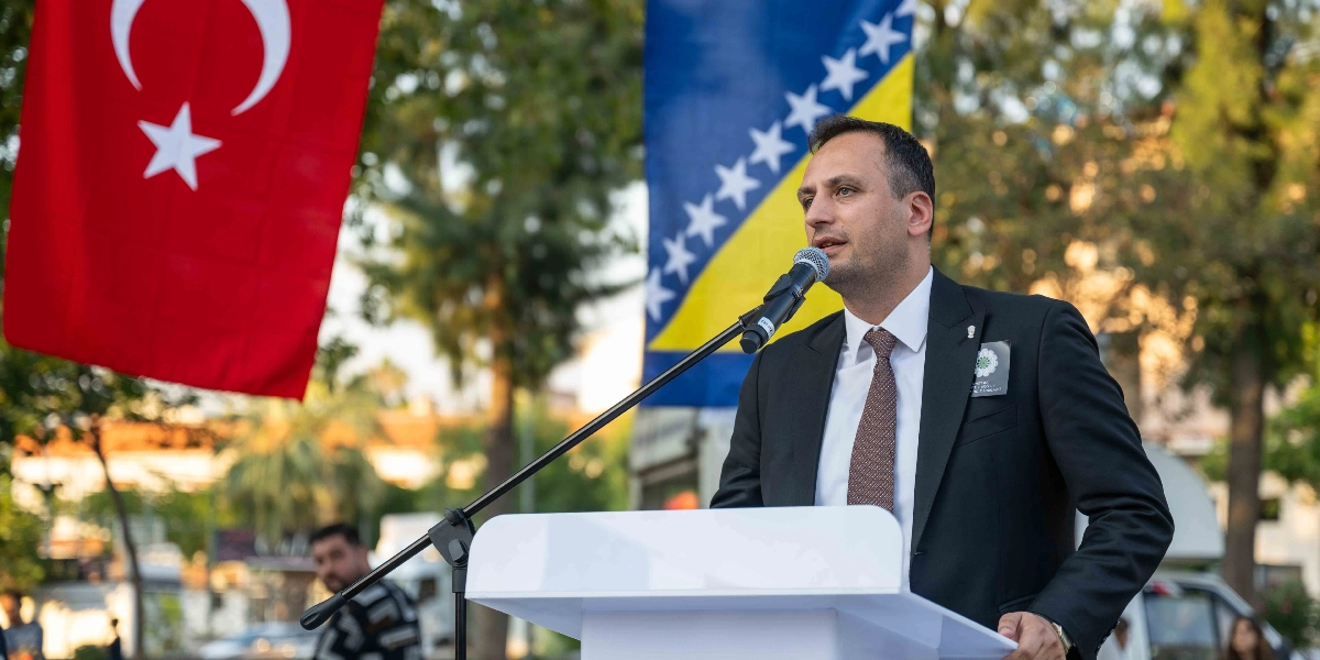 Koyu renk takım elbiseli ve kravatlı bir erkek, açık havada beyaz bir kürsü arkasında mikrofonla konuşma yapıyor. Konuşmacının arkasında sol tarafta Türk bayrağı, sağ tarafta ise Bosna-Hersek bayrağı asılı duruyor. Konuşmacının ceketinin yakasında Srebrenica soykırımını simgeleyen 