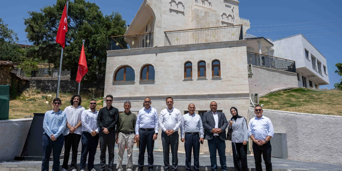 Taş mimariye sahip, balkonlu ve kemerli pencereleri olan modern bir binanın önünde yan yana duran, çoğunluğu erkeklerden oluşan on kişilik bir grup. Grubun sol tarafında, direklerde asılı iki Türk bayrağı ve yanında Atatürk büstü yer alıyor. Açık havada, güneşli bir günde çekilmiş bu karede, gruptaki kişiler resmi ve yarı resmi kıyafetlerle kameraya poz veriyor.