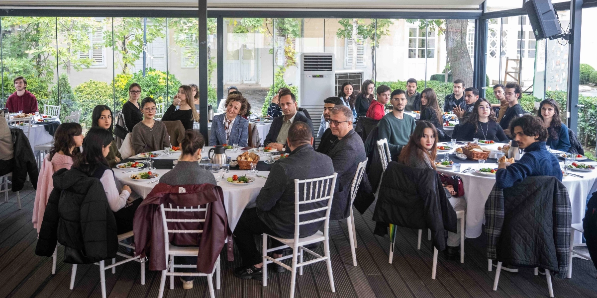 Camla çevrili aydınlık bir mekânda, beyaz örtülü yuvarlak masaların etrafında toplanmış, çoğunluğu gençlerden oluşan bir grup insan bir arada oturuyor. Masalarda tabaklar, bardaklar ve yiyecekler bulunuyor. Katılımcılar sohbet ederek yemek yiyor, bazıları kameraya bakarken bazıları yanındaki kişilerle konuşuyor. Mekânın arka planında ağaçlıklı bir bahçe manzarası ve beyaz bir klima ünitesi görülüyor.