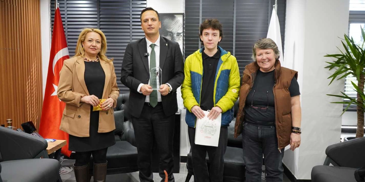 Bornova Belediye Başkanı Mustafa İduğ, yanında bir kadın ve bir gençle birlikte duruyor. Başkan İduğ, elinde şeffaf bir plaket tutarken, yanındaki genç üzerinde Bornova Belediyesi logosu bulunan beyaz bir hediye çantası taşıyor. Arka planda Türk bayrağı, siyah deri koltuklar ve ofis dekorasyonu yer alıyor.