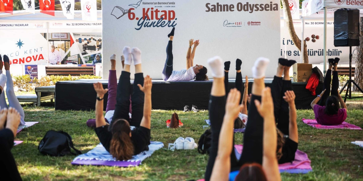 Bornova Belediyesi 6. Kitap Günleri kapsamında, Sahne Odysseia önündeki çim alanda yoga veya pilates yapan bir grup insan. Katılımcılar matlarının üzerinde sırtüstü uzanmış, bacaklarını ve kollarını havaya kaldırmış egzersiz yaparken, sahnedeki eğitmen de aynı hareketi gösteriyor. Arka planda etkinlik logosunun yer aldığı büyük bir sahne panosu ve stantlar bulunuyor.