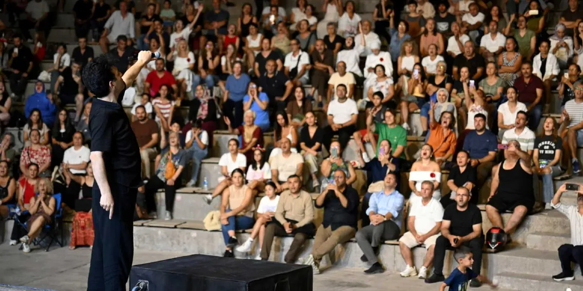 Açık hava tiyatrosunda sahneye sırtı dönük şekilde elinde mikrofonla konuşan siyah kıyafetli bir sanatçı ve onu izleyen, basamaklı oturma düzenine yerleşmiş kalabalık bir seyirci grubu. Bazı seyirciler ellerindeki telefonlarla çekim yaparken, bazıları alkışlıyor veya sahneye doğru bakıyor. Ön planda küçük bir çocuk sahneye doğru yürüyor.