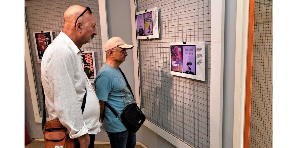 Beyaz gömlekli, kel bir adam ile bej şapkalı ve mavi tişörtlü bir adam, bir sergi alanındaki tel panoya asılmış Orhan Kemal'in 