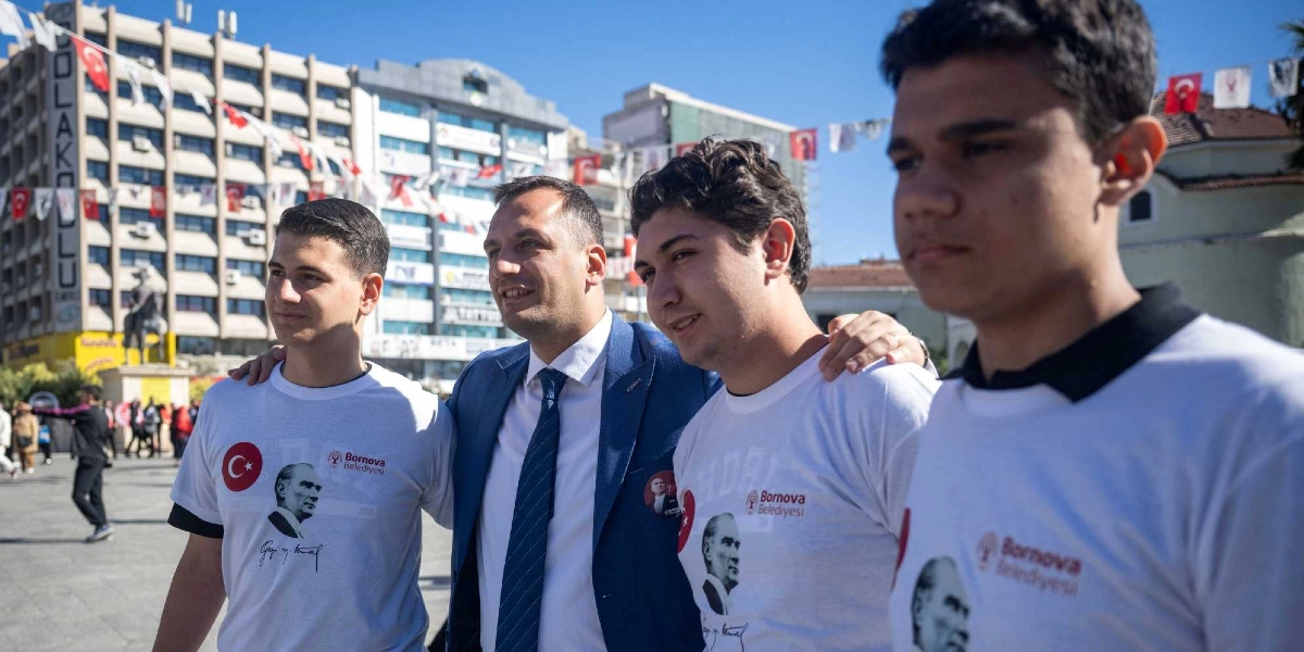 Bornova Belediye Başkanı, üzerinde Atatürk portresi ve Bornova Belediyesi logosu bulunan beyaz tişörtler giymiş üç gençle birlikte açık havada yan yana durarak gülümsüyor. Arka planda Türk bayraklarıyla süslenmiş bir meydan ve binalar yer alıyor.