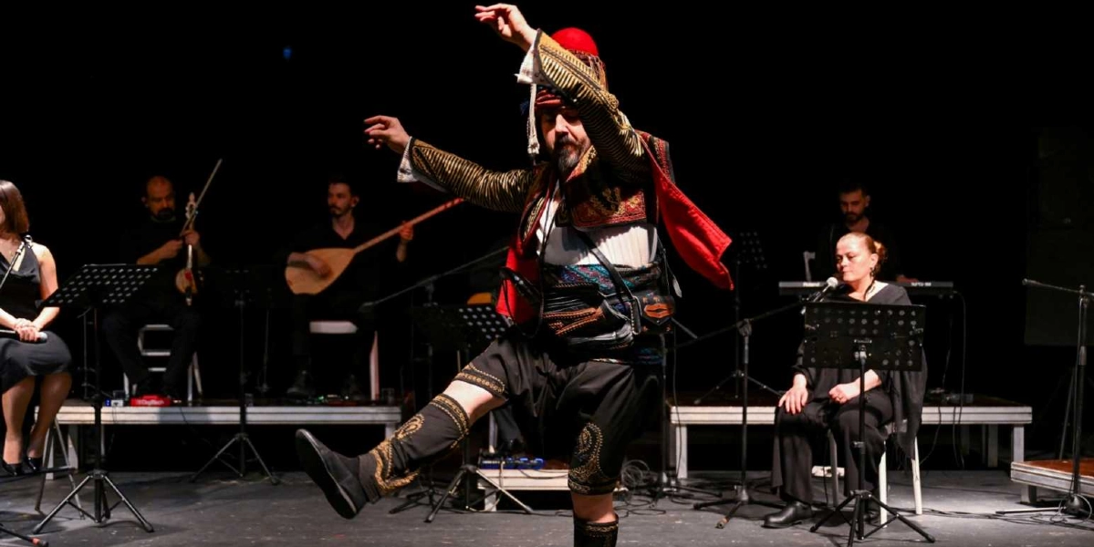 Siyah bir sahnede, geleneksel Ege zeybek kıyafetleri giymiş bir erkek dansçı, bir ayağını havaya kaldırmış figür sergiliyor. Dansçı, işlemeli ceket, kırmızı fes ve şalvar giyiyor. Arka planda ise müzisyenler oturuyor; solda keman ve bağlama çalanlar, sağda ise mikrofon başında şarkı söyleyen bir kadın yer alıyor.