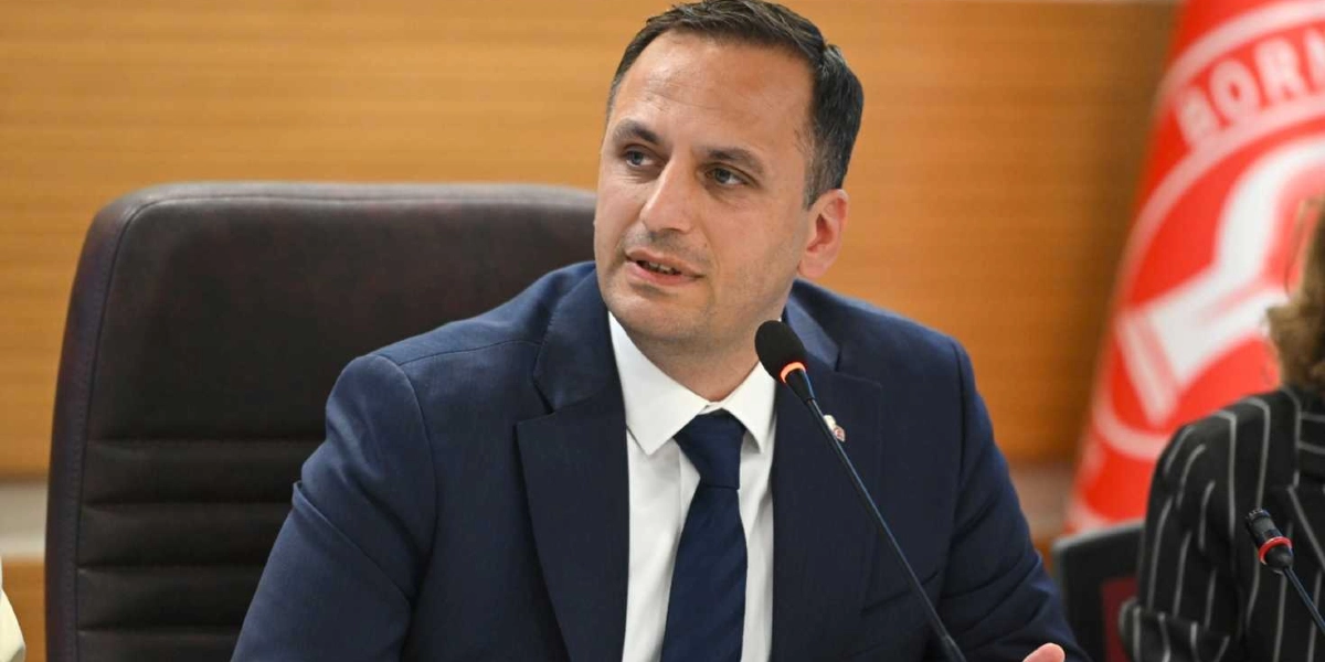 Koyu renk ceket ve lacivert kravat giymiş, önündeki mikrofona konuşan bir erkek yönetici, toplantı masasında otururken görülüyor. Arka planda Bornova Belediyesi'ne ait olduğu anlaşılan kırmızı renkli bir bayrak yer alıyor.