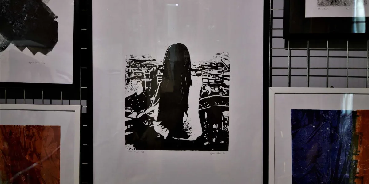 Bir sergi duvarında asılı duran, siyah beyaz baskı tekniğiyle yapılmış bir sanat eseri. Eserde, arkası dönük, uzun saçlı bir kadın figürü, yüksek bir noktadan aşağıya doğru uzanan şehir manzarasına bakarken tasvir ediliyor. Çerçevenin alt kısmında sanatçı imzası ve eserin bilgileri yer alıyor.