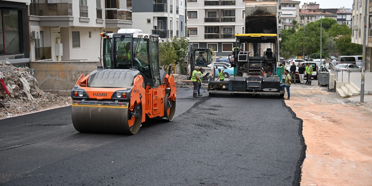 Şehir içindeki bir sokakta asfalt serme çalışması yapılıyor. Ön planda turuncu renkli bir Hamm silindir, hemen arkasında ise Vögele marka bir asfalt serici makine bulunuyor. İşçiler yelekleriyle çalışma alanında görev alırken, yeni dökülen pürüzsüz siyah asfalt yolun bir kısmını kaplıyor. Çevrede çok katlı apartmanlar ve park halindeki araçlar görülüyor.