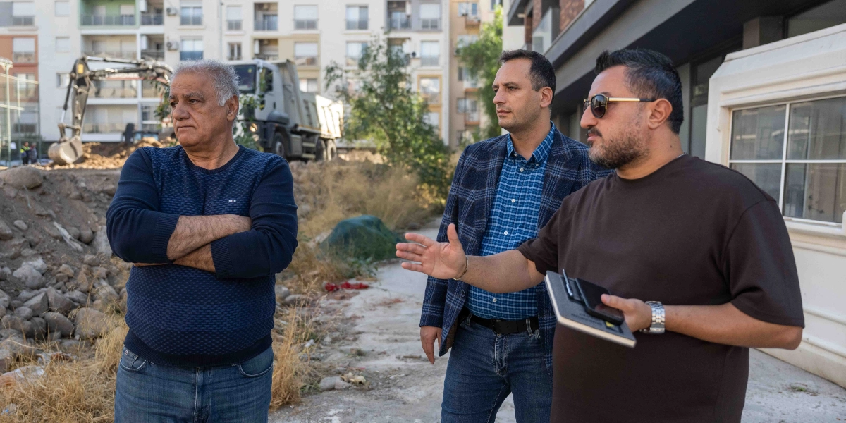 Üç erkek, arka planda iş makinelerinin ve inşaat halindeki bir alanın bulunduğu bir arazide incelemelerde bulunuyor. En solda kolları kavuşmuş şekilde duran gri saçlı bir adam, ortada ekose ceketli bir adam ve en sağda elinde not defteri tutarak eliyle alanı işaret eden güneş gözlüklü bir adam yer alıyor.