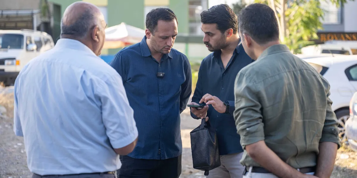 Açık havada, bir otopark alanında bir araya gelmiş dört erkek, ellerindeki akıllı telefona bakarak bir konu üzerinde tartışıyor. Gruptaki iki kişi telefon ekranını incelerken, diğer ikisi onları dikkatle dinliyor. Arka planda park halindeki araçlar ve binalar görülüyor.