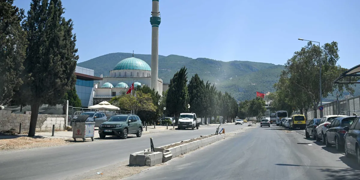 Geniş bir cadde üzerinde, yol kenarına park etmiş araçların bulunduğu ve orta refüjünde inşaat çalışmaları süren Pınar Caddesi'nin görünümü. Arka planda yeşil kubbeli ve minareli bir cami ile yüksek dağlar yer alıyor. Yolun sol tarafında ağaçlar, sağ tarafında ise sıralı park etmiş otomobiller ve ağaçlık bir alan bulunuyor. Gökyüzü açık ve güneşli.