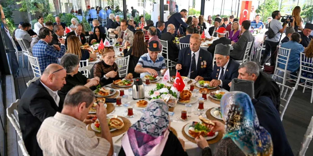 Gaziler Günü vesilesiyle düzenlenen bir yemekte, yuvarlak masalar etrafında toplanmış gaziler ve yakınları bir arada yemek yiyor. Ön plandaki masada, üzerinde Türk bayrağı bulunan bir kahvaltı sofrasında oturan katılımcılar, çaylarını içip sohbet ediyor. Arka planda ise benzer şekilde düzenlenmiş diğer masalarda çok sayıda davetli yer alıyor. Mekan, cam duvarlarla çevrili, aydınlık ve geniş bir salon görünümünde.