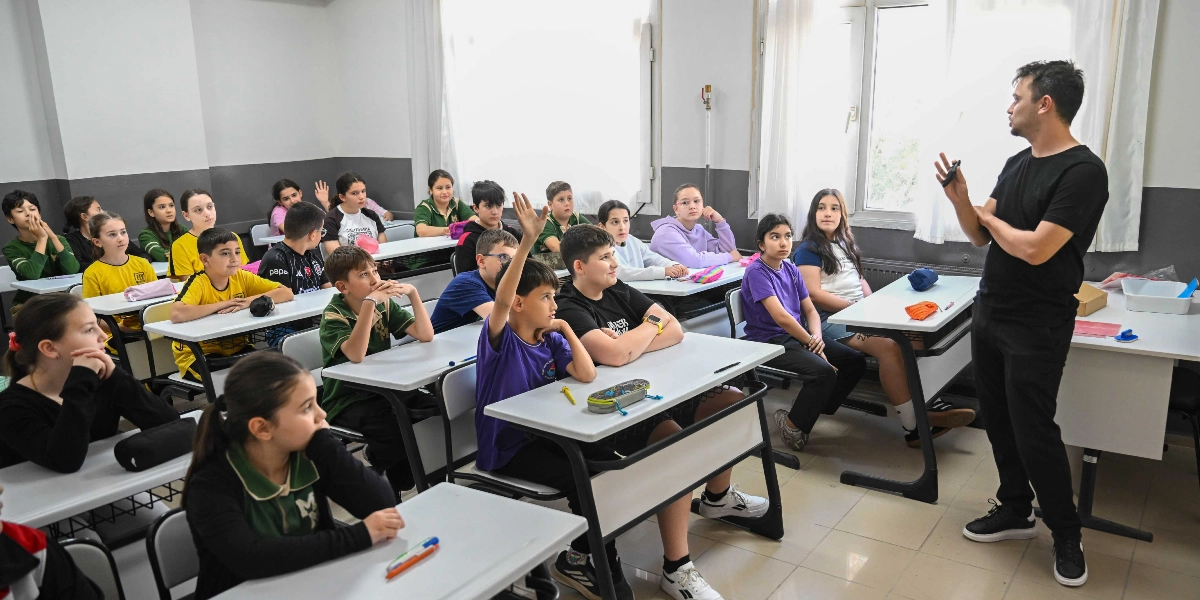 Sınıf ortamında, siyah tişörtlü bir eğitmen ayakta öğrencilere bir şeyler anlatırken, sıralarda oturan ortaokul çağındaki öğrenciler onu dikkatle dinliyor ve içlerinden biri söz almak için elini kaldırmış durumda.