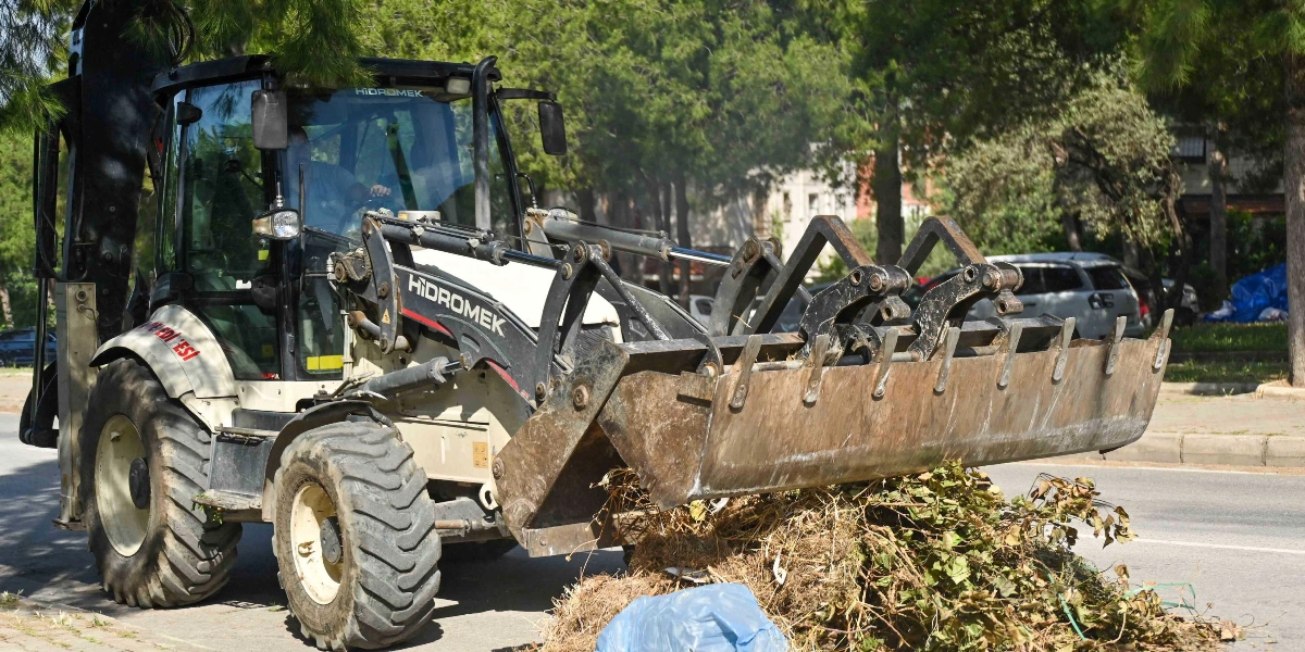 Bornova Belediyesi'ne ait Hidromek marka bir iş makinesi, yol kenarındaki kurumuş otları ve çalıları temizleyerek kepçesiyle topluyor.